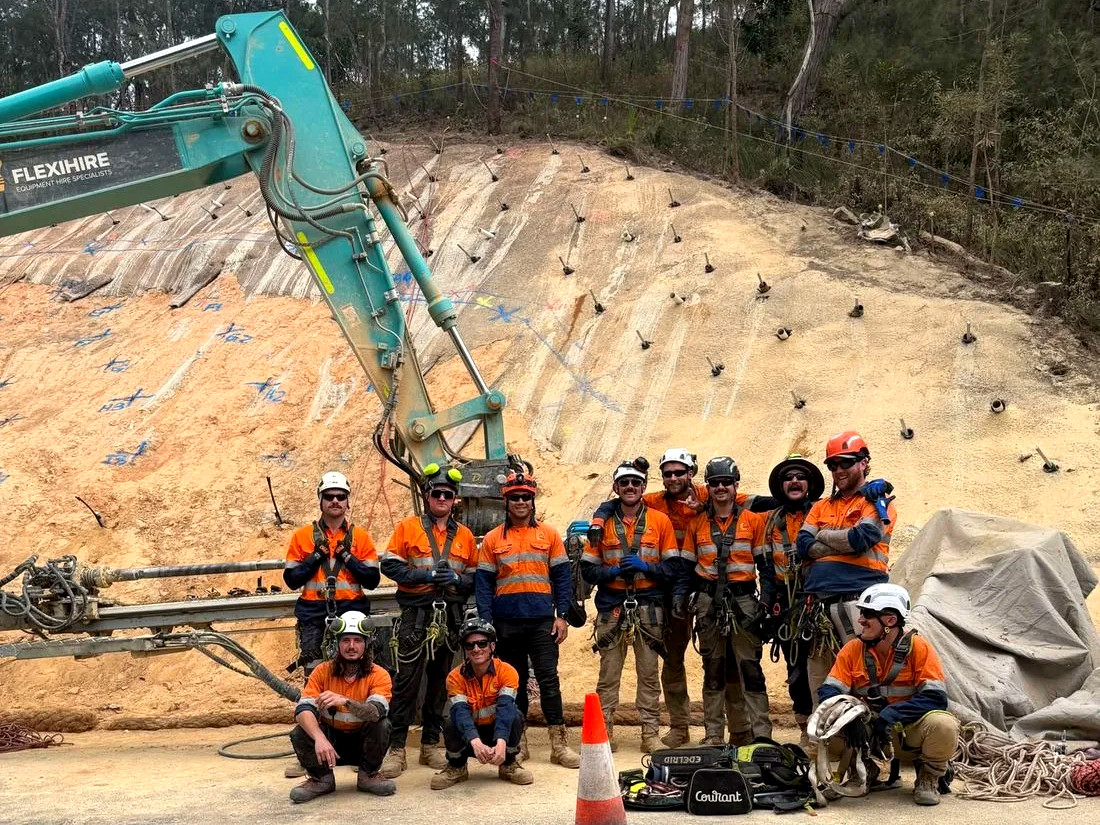 A Pointe slope crew of twelve in front of a soil nailed rock face, with an excavator mounted drill rig
